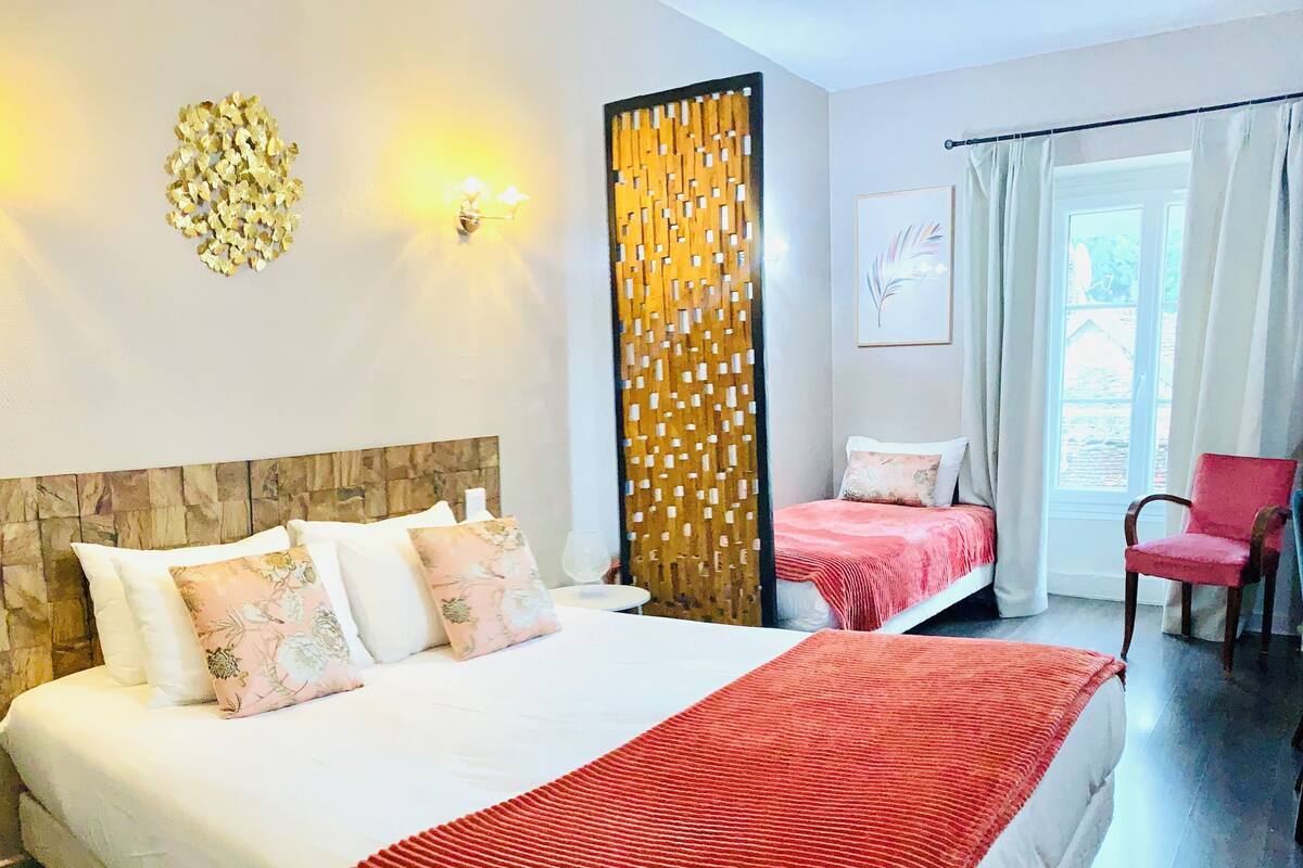 A hotel room with a large bed, a smaller bed, a red throw blanket, a wooden headboard, a decorative wall panel, a framed picture, and a red chair.