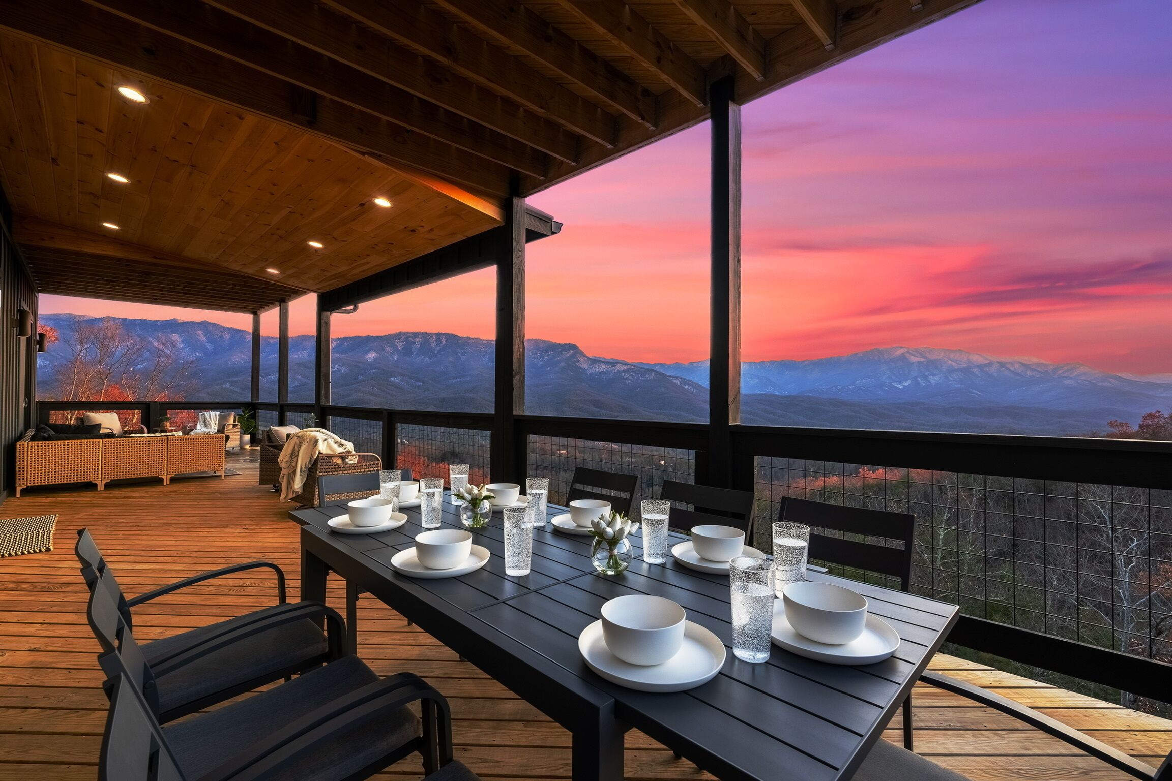 A wooden deck with a table set for dining, overlooking a mountainous landscape during sunset.