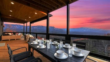 A wooden deck with a table set for dining, overlooking a mountainous landscape during sunset.