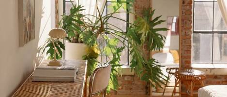 A cozy room with a brick wall, a wooden herringbone chair, a small round table, a bed, and a large potted plant.