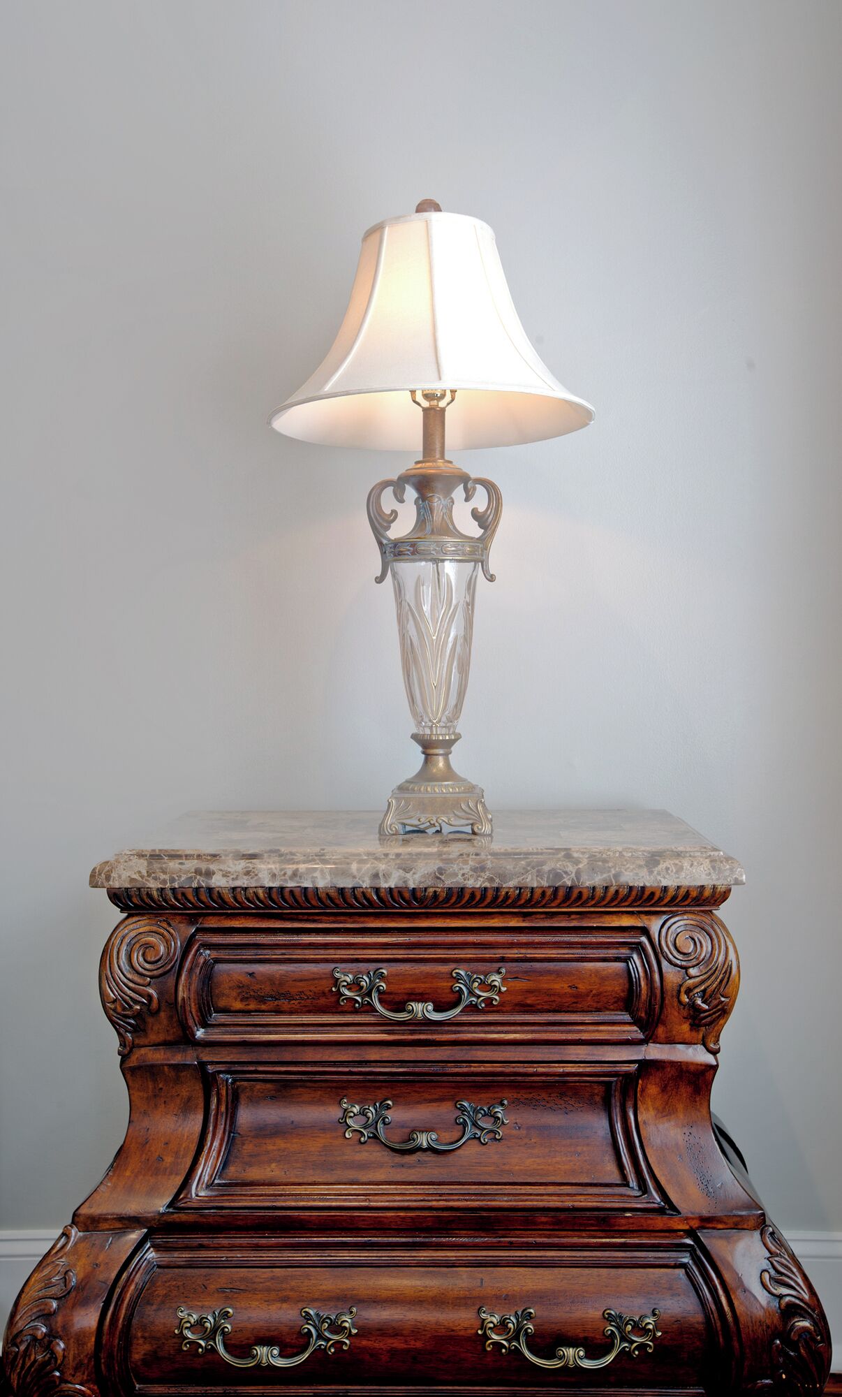 A table with a lamp on top, featuring a marble top and ornate wooden base with drawers.