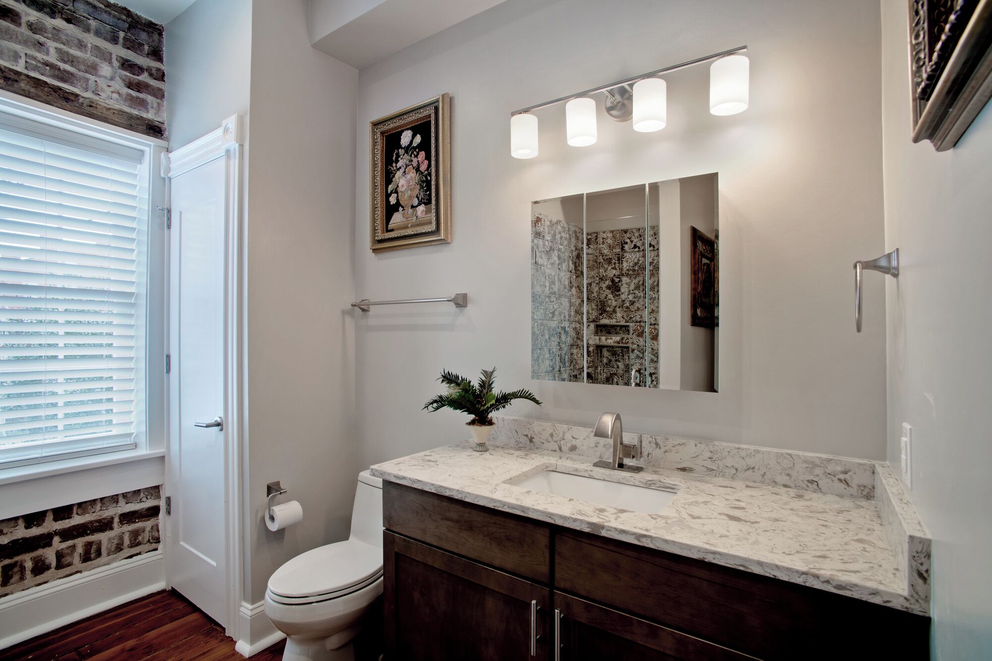 A bathroom with a white toilet, a marble sink, a mirror with lights, a potted plant, and a brick wall.