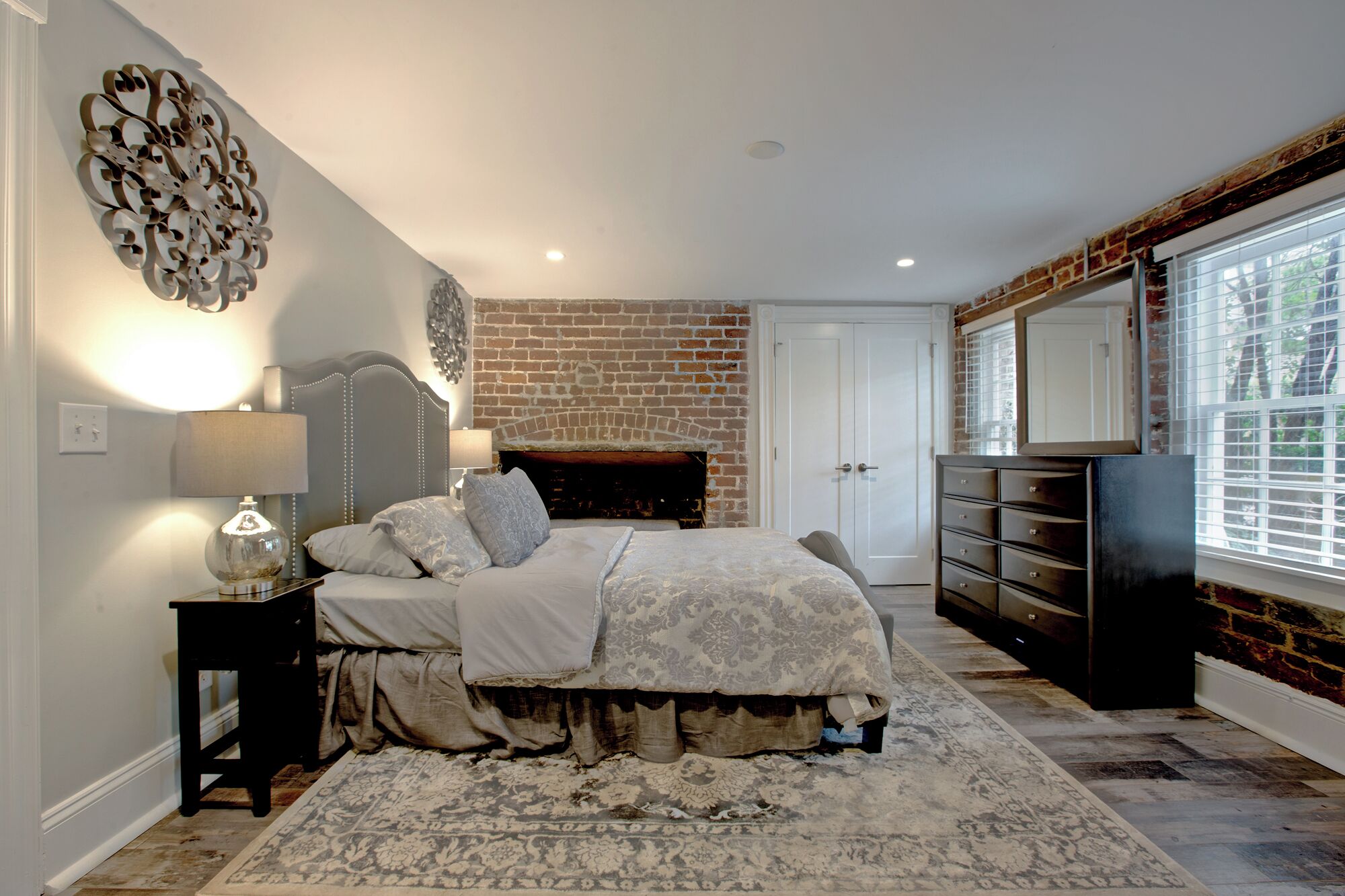 A bedroom with a brick fireplace, a bed with a patterned bedspread, a dresser, and a large mirror.