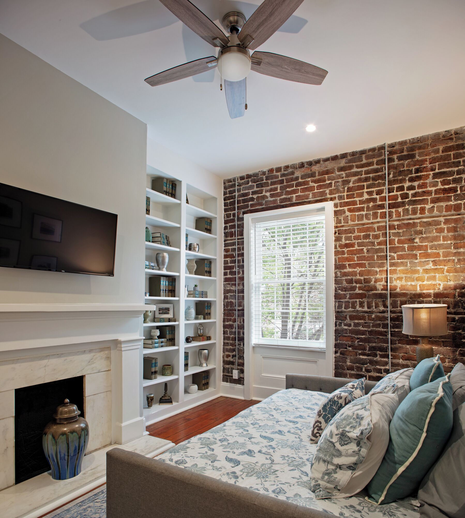 A bedroom with a brick wall, a fireplace, a TV mounted above it, and a bed with patterned bedding.