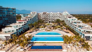 A resort with multiple buildings, a central swimming pool, and a beach area with sun loungers and palm trees. - Luxury Beachfront Apartment with Infinity Swimming Pools in Nuevo Vallarta (Nuevo Vallarta)