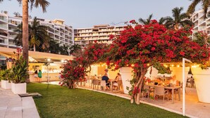 An outdoor dining area with tables and chairs, surrounded by lush greenery and flowering plants. - Luxury Beachfront Apartment with Infinity Swimming Pools in Nuevo Vallarta (Nuevo Vallarta)