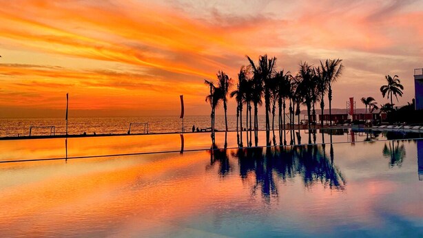 A sunset over a calm pool with palm trees and a distant view of the ocean. - Luxury Beachfront Apartment with Infinity Swimming Pools in Nuevo Vallarta (Nuevo Vallarta)