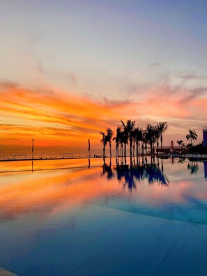 A sunset over a calm pool with palm trees and a distant view of the ocean. - Luxury Beachfront Apartment with Infinity Swimming Pools in Nuevo Vallarta (Nuevo Vallarta)