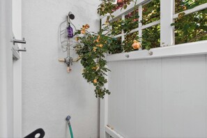 Een witte gevel met een raam, een metalen slangkastel met een paarse fles, en een hangplant met gele bloemen.