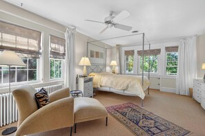 A bedroom with a four-poster bed, a ceiling fan, a bedside table with lamps, a chair, a rug, and large windows with curtains.