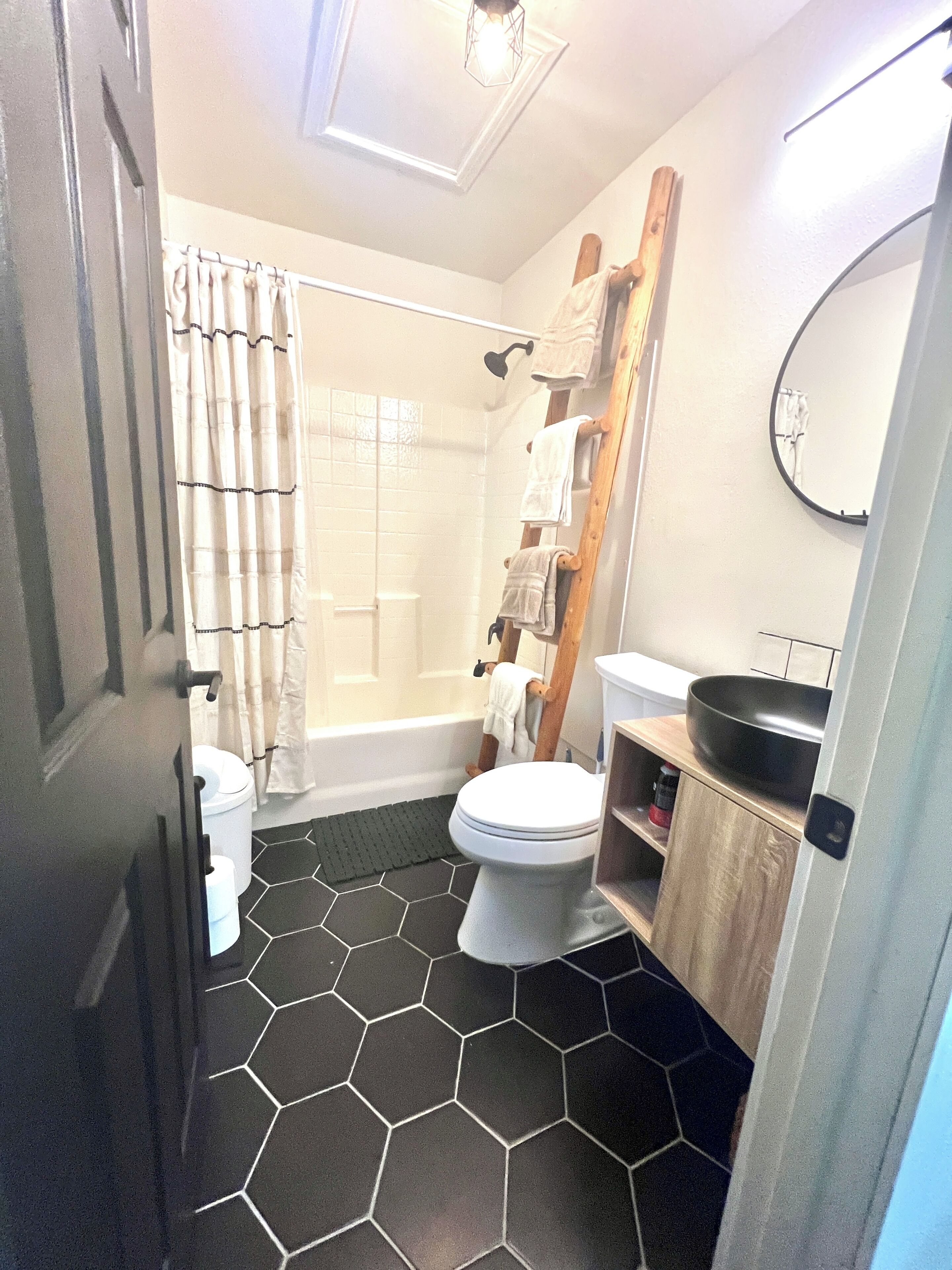 Et badeværelse med gulv af sekskantede fliser, toilet, brusebad og vask med en træhylde.