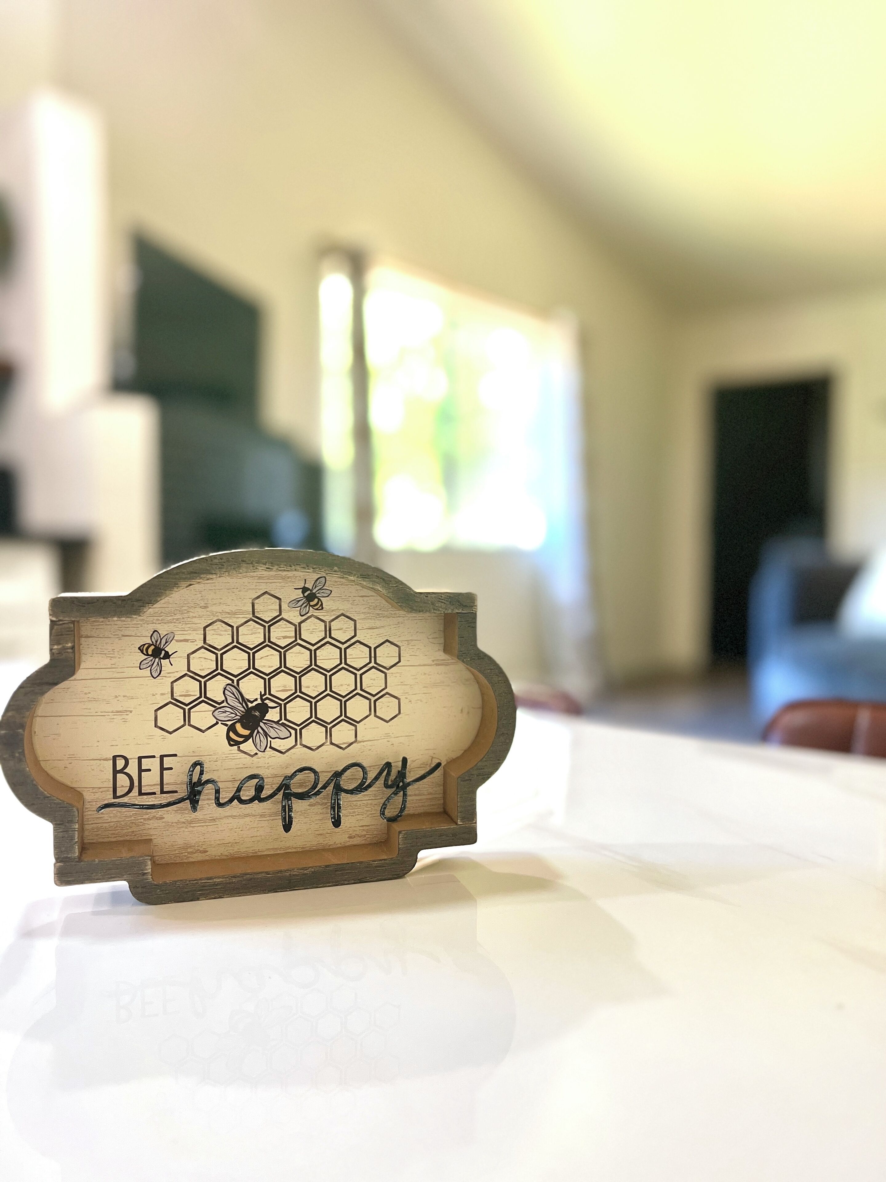 A wooden sign with a honeycomb design and bees, placed on a table in a room with a blurred background.