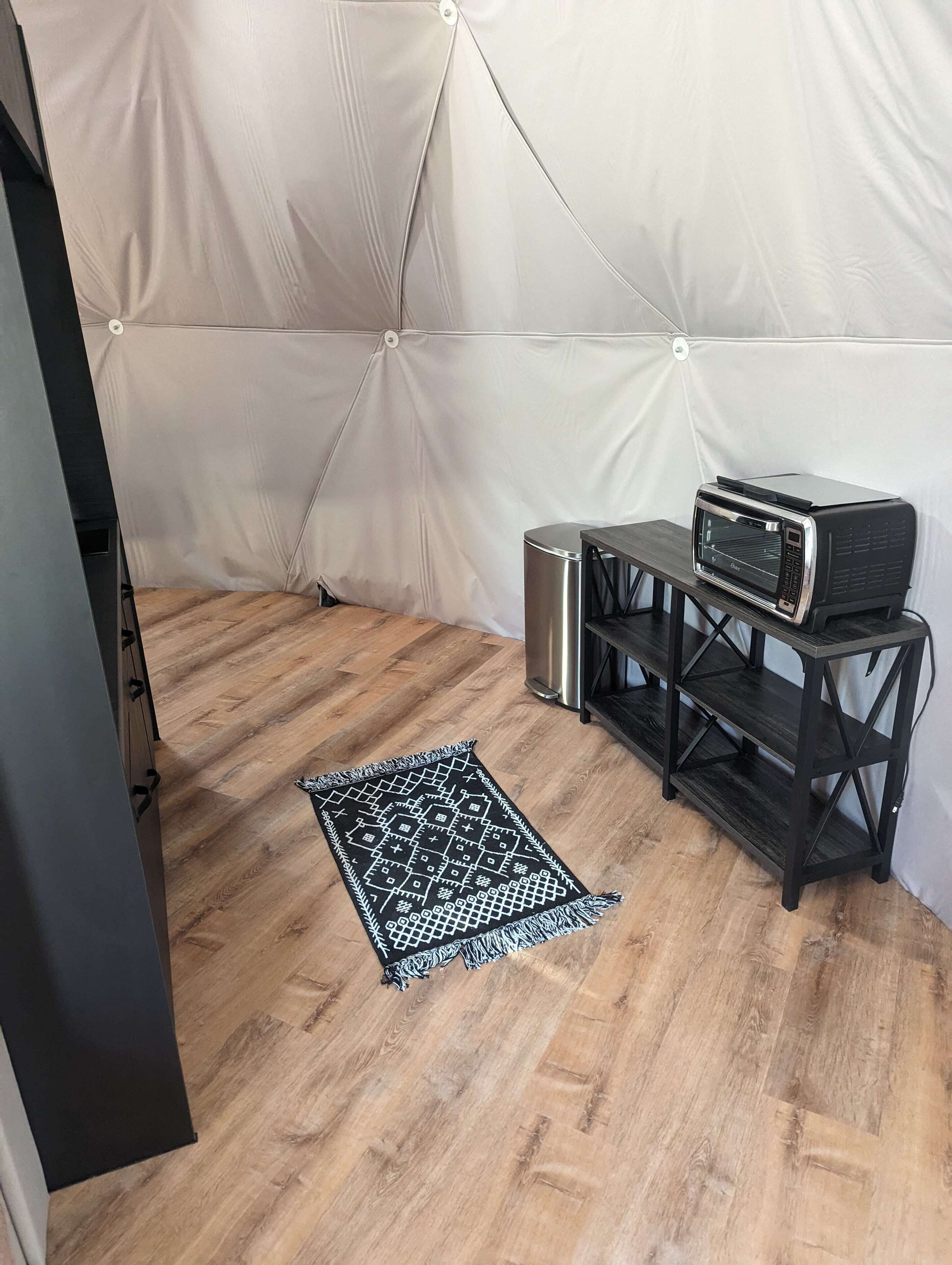 A tented room with a microwave on a shelf, a trash bin, and a patterned rug on the floor.
