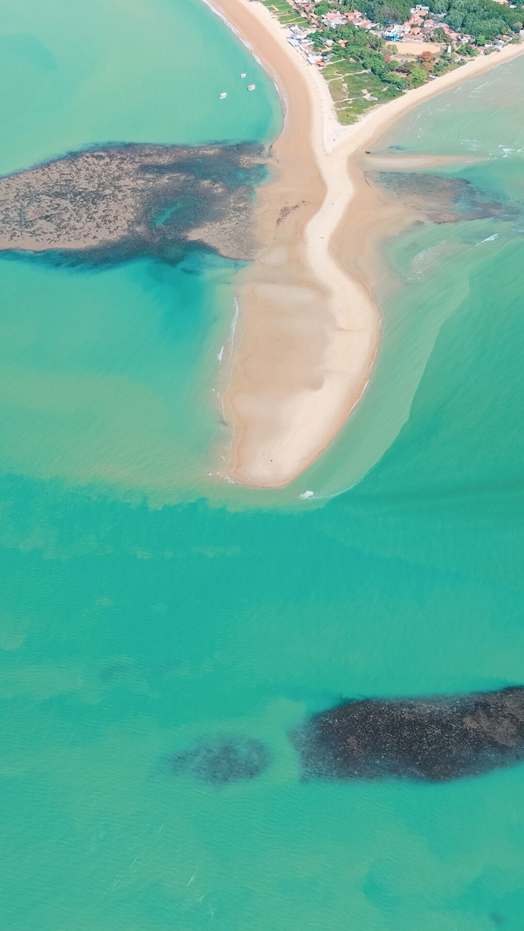 An aerial view of a beach with clear turquoise waters, a sandy shoreline, and a small settlement in the background.