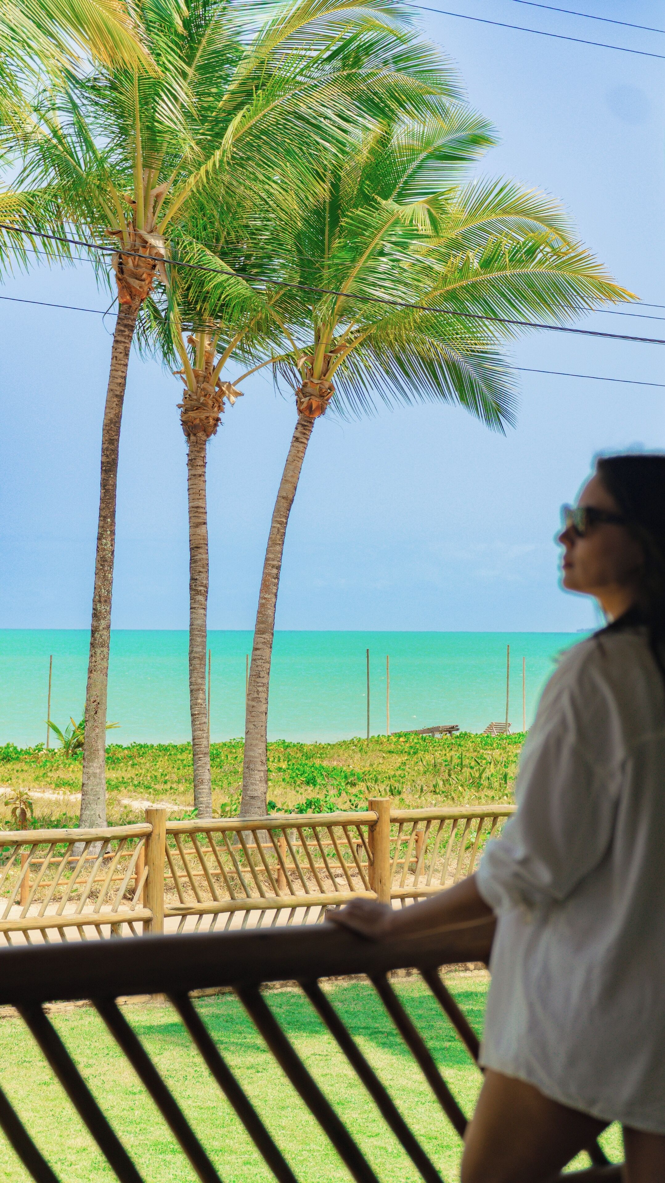 Iemand staat op een balkon met uitzicht op een strand, palmbomen en een heldere lucht.