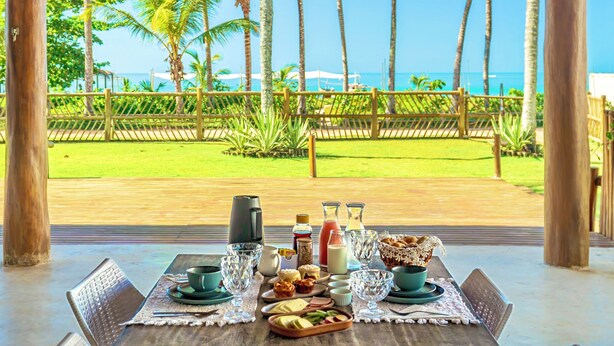 A dining table set with breakfast items, a view of the ocean, and palm trees. - Casa Mandala Corumbau (Corumbau)