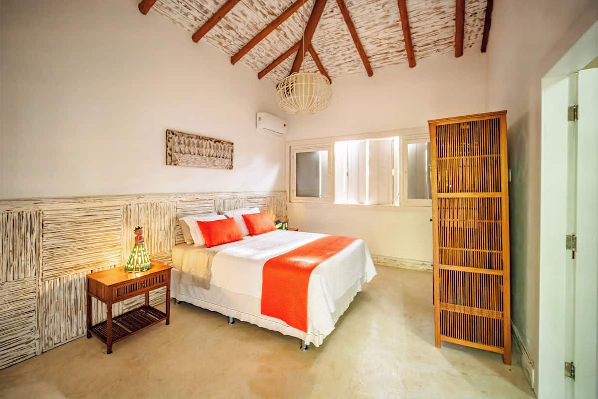 A bedroom with a bed, a nightstand, a wall-mounted lamp, a wooden wardrobe, and a window with blinds.