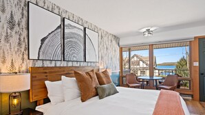 Habitación de hotel con una cama grande, cabecera, mesita de noche con lámpara y vistas al exterior a través de amplias ventanas.