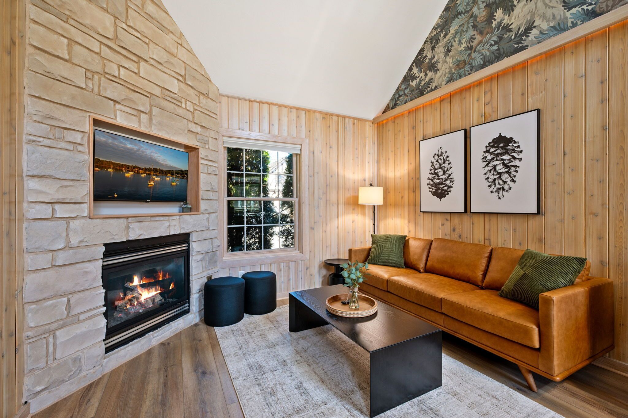 Un salon chaleureux avec une cheminée en pierre, des murs en bois, un canapé en cuir et des œuvres d’art encadrées.