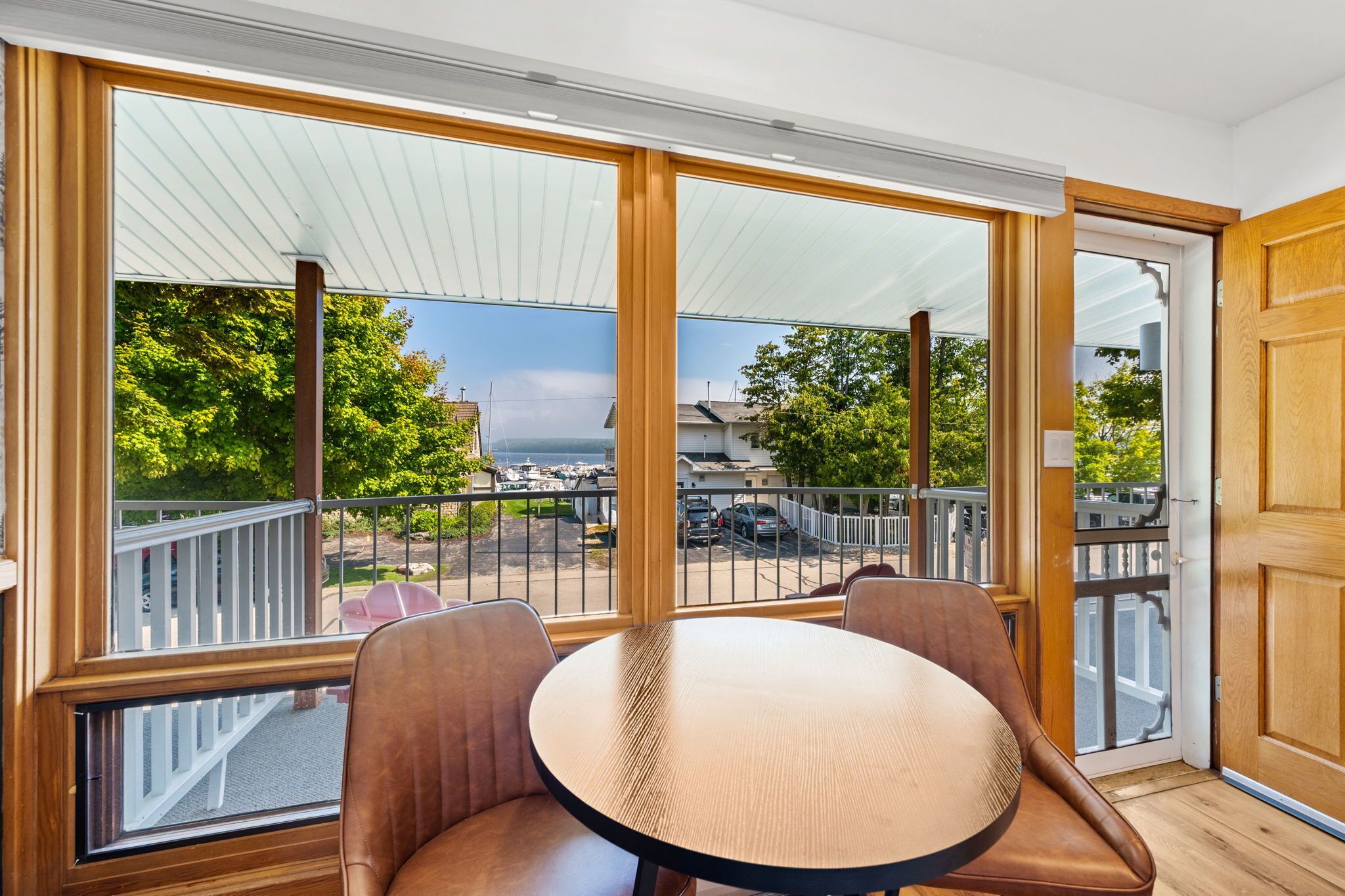 A room with a large sliding glass door leading to a balcony with a view of a marina and boats. There is a round table with two chairs in the room.