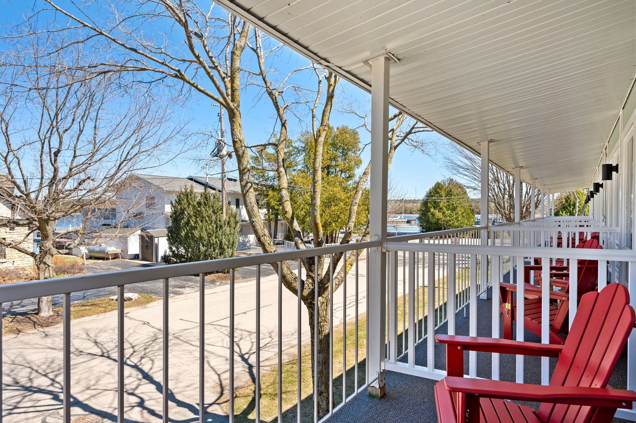 A covered porch with red Adirondack chairs, a metal railing, and a view of a residential street with trees and houses.