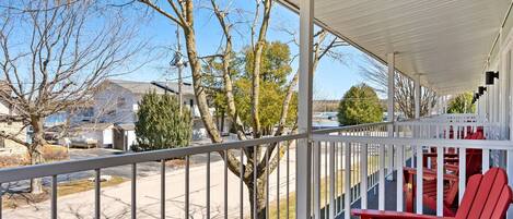 A covered porch with red Adirondack chairs, a metal railing, and a view of a residential street with trees and houses.