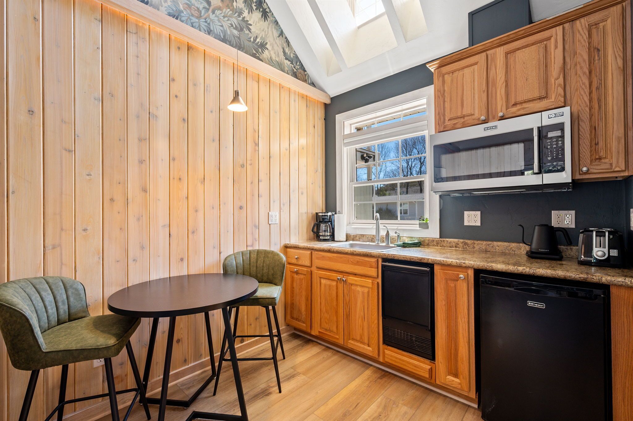 A compact kitchen with wooden cabinets, a microwave, a dishwasher, and a small dining area with two chairs.