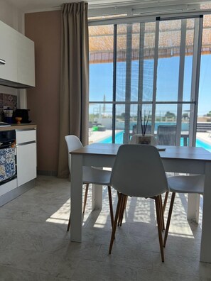 Eine moderne Küche mit weißen Schränken und einem Essbereich inklusive Tisch und Stühlen. Ein großes Fenster bietet Blick auf einen Pool und ein Gebäude im Hintergrund.