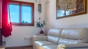 A living room with a beige leather sofa, a window with a red curtain, a wall-mounted wind chime, a small table with a vase, and a framed abstract painting.