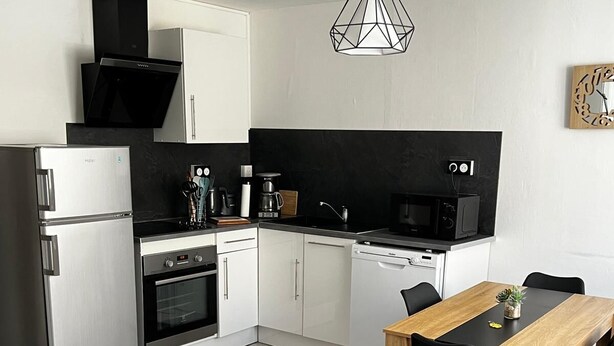 Una cocina moderna con armarios blancos, un refrigerador de acero inoxidable, un horno empotrado, un microondas y una cafetera. En primer plano, se aprecia una mesa de comedor de madera con sillas negras y un sofá gris.