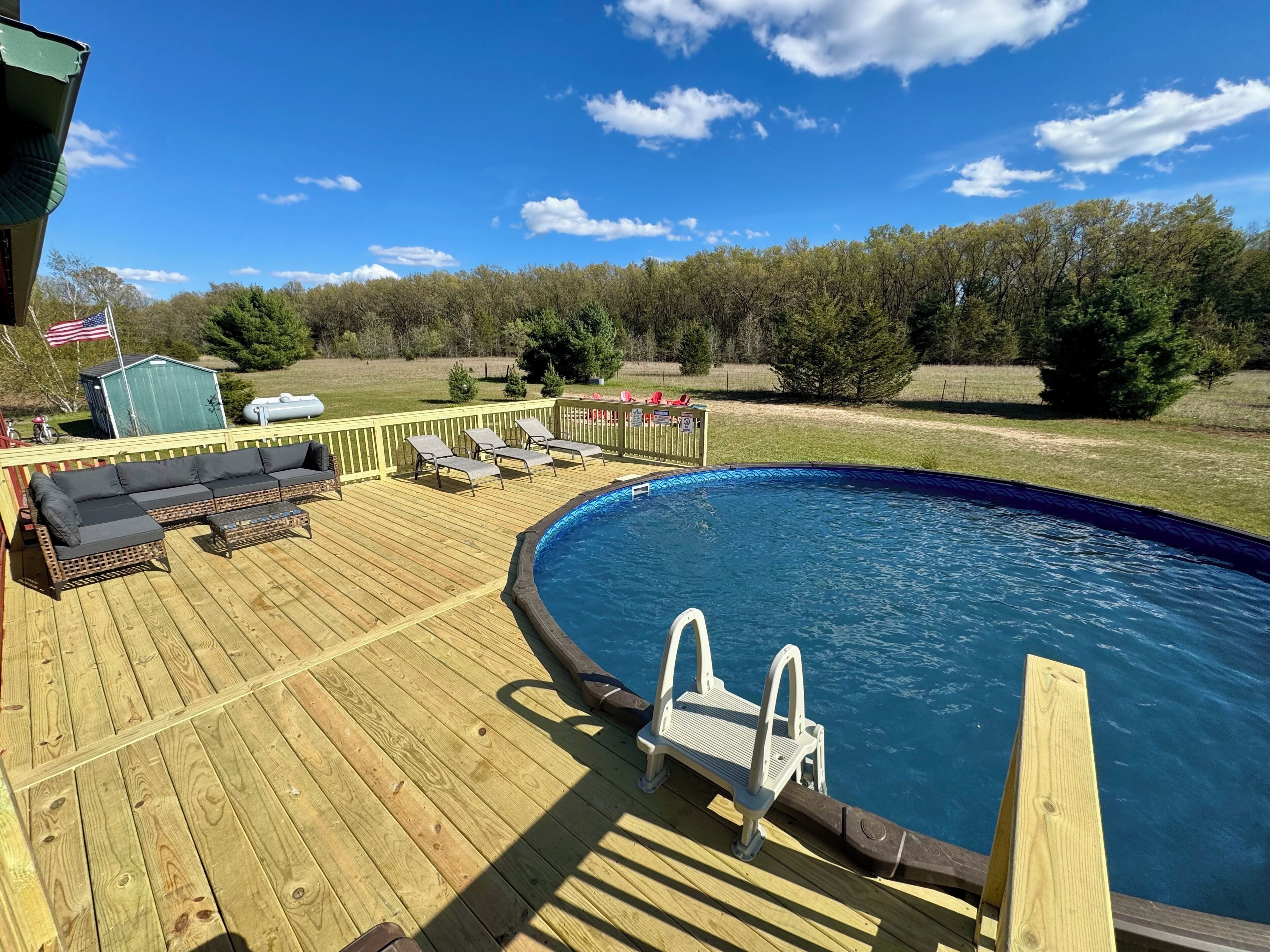 A wooden deck with a pool, lounge chairs, and a small shed with an American flag.