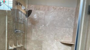 A shower with a glass door, beige tiles, and a shelf with toiletries. - Beautiful,comfortable, safe suite with private entrance. (Dallas)