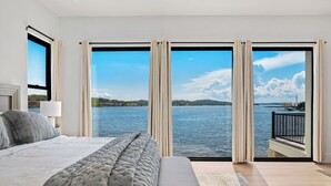 Quarto com cama grande, um banco e vista para um corpo d'água através de janelas do chão ao teto.