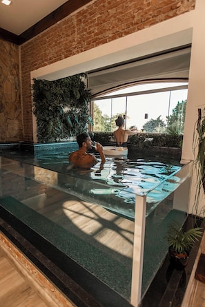 A man in a glass-bottomed indoor pool with a brick wall and a large window.