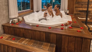 A couple in a hot tub with rose petals, wooden beams, and a view of trees. - Roteiros Adorai (Piedade)
