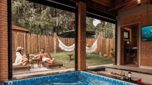 An indoor swimming pool with a wooden ceiling, a brick wall, and a view of a backyard with a hammock. - Roteiros Adorai (Piedade)