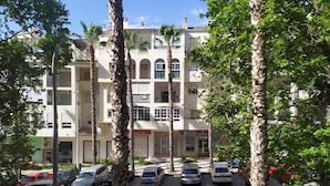 A multi-story residential building with balconies, parked cars, and palm trees.