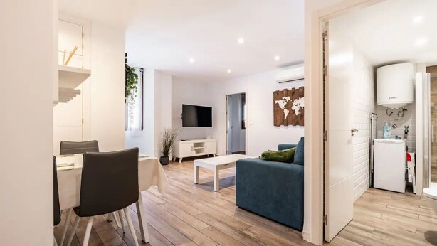 A modern living room with a sofa, coffee table, and TV. There is a dining area with chairs and a table. The room has wooden flooring and white walls.