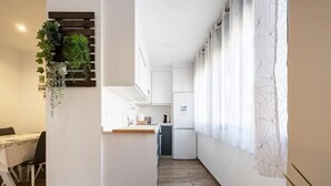 A narrow kitchen with white cabinets, a wooden countertop, and a small refrigerator. There is a dining area with a table and chairs to the left.