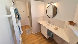 A modern bathroom with a white vanity, a round mirror, and a wooden floor. - Apartment n°1 : Sybèle (La Gaubretière)