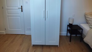 Ein Zimmer mit einem weißen Schrank, einem kleinen Tisch mit Lampe und einem Bett mit gestreiftem Kissen.