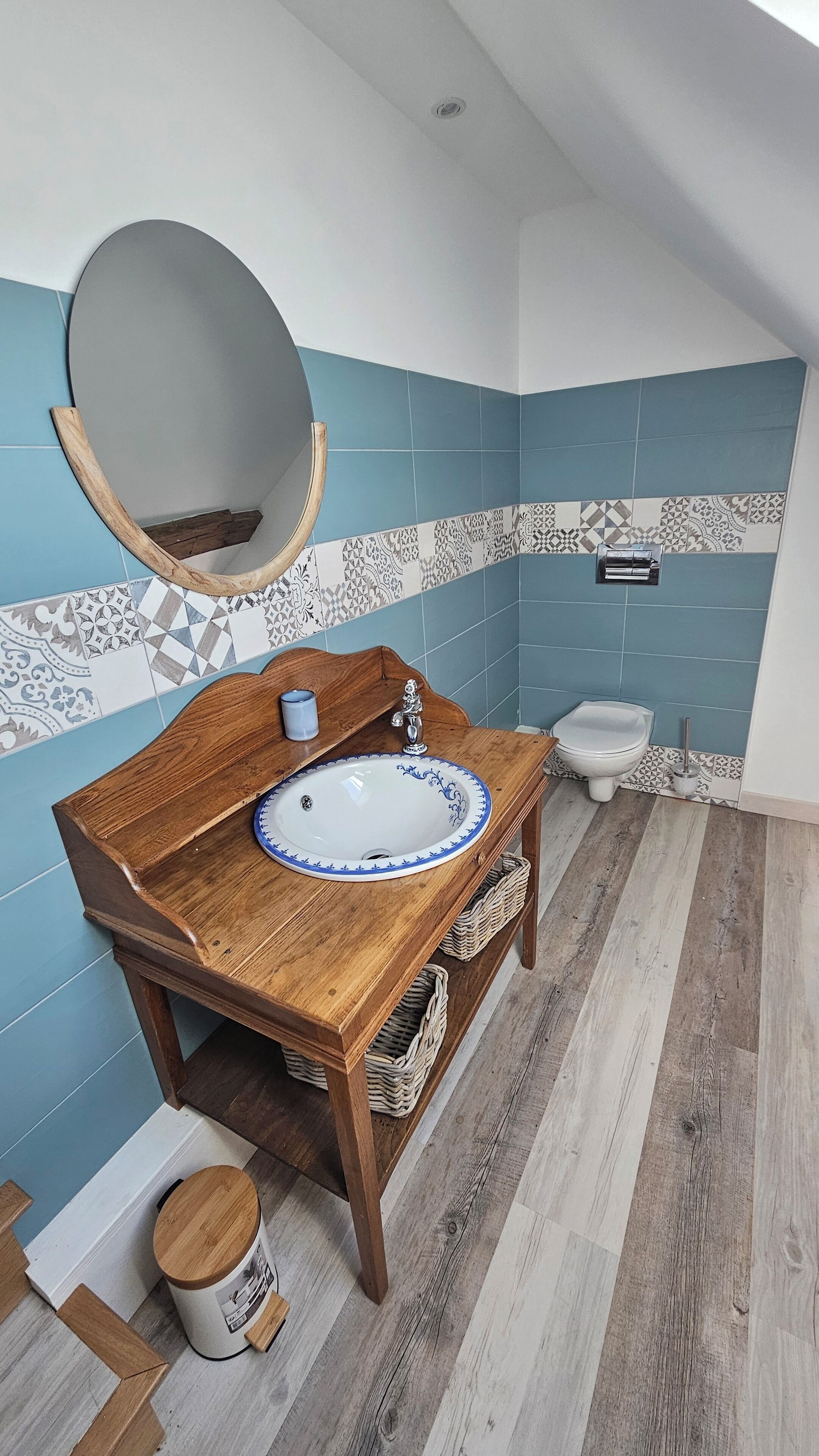 A bathroom with a wooden vanity, a round mirror, a white sink with blue trim, and a toilet. The walls are tiled in blue and white patterns.