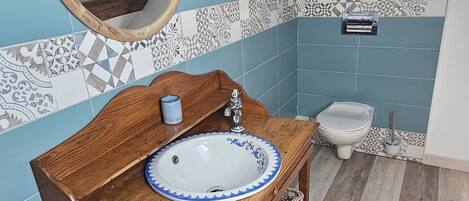 A bathroom with a wooden vanity, a round mirror, a white sink with blue trim, and a toilet. The walls are tiled in blue and white patterns.