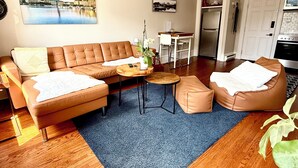 A living room with a sofa, ottoman, coffee table, and a small side table with a plant. There is a kitchen area with a refrigerator and a dining table.