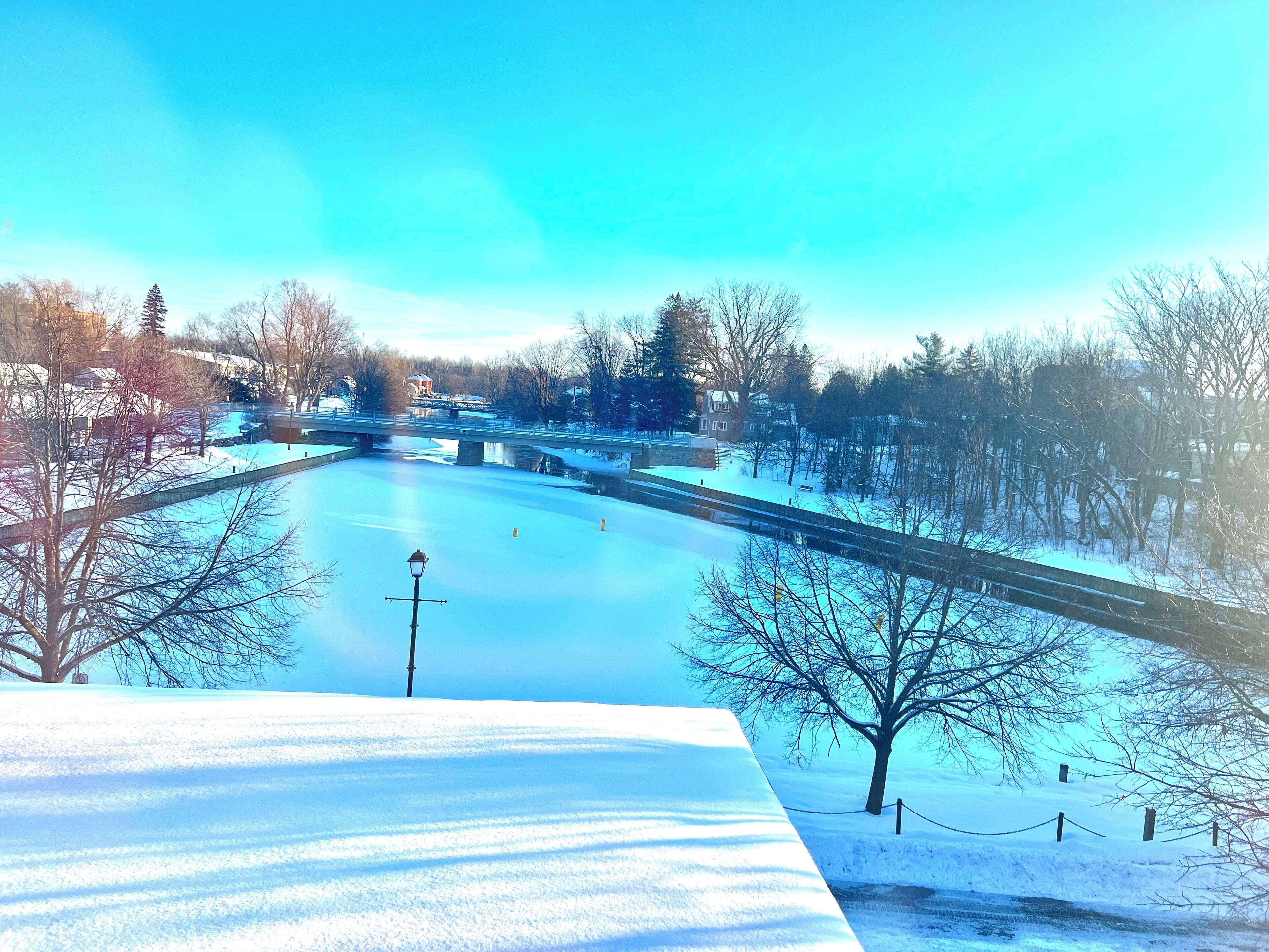 A snow-covered river with a bridge, trees, and a lamppost.