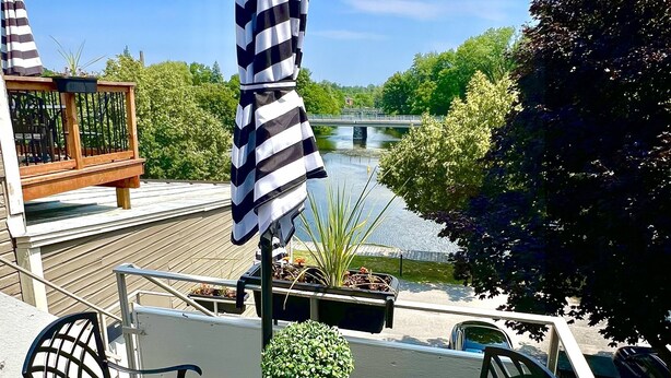 A deck with a round table and chairs, a striped umbrella, and a view of a river and greenery.