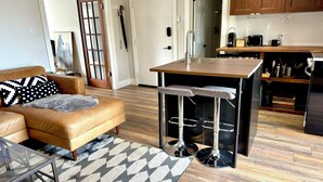 A modern living room with a leather sofa, a glass coffee table, a kitchen area with wooden cabinets, a bar stool, and a patterned rug.