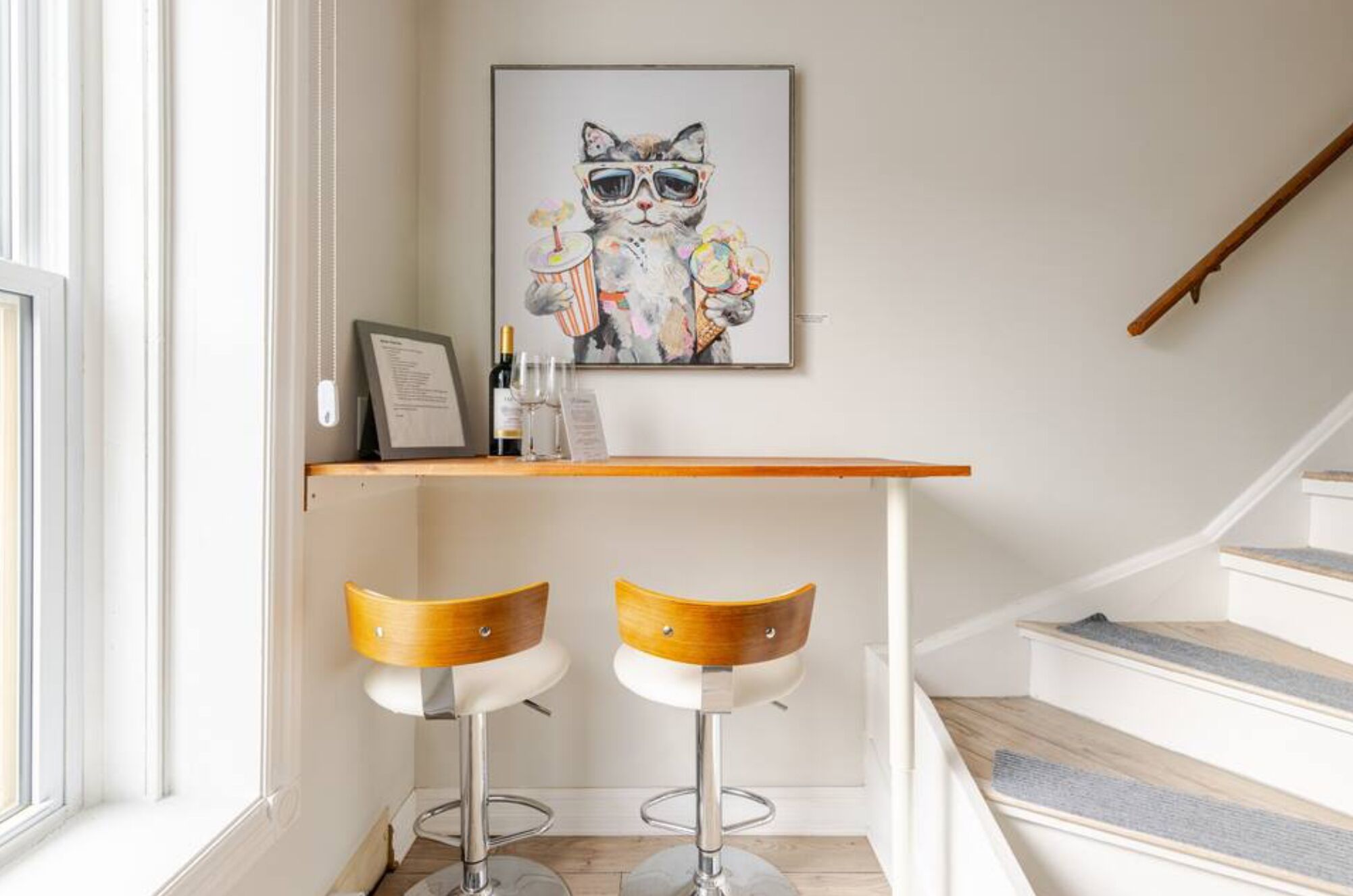En modern inredning med en trappa, två barstolar, ett inramat konstverk som föreställer en katt och en liten bardisk med glas och en meny.