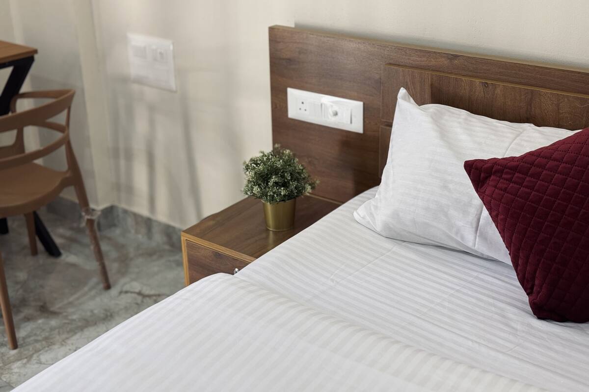 Een netjes opgemaakt bed met houten hoofdbord, een klein nachtkastje met een plant, en een stoel op de achtergrond.