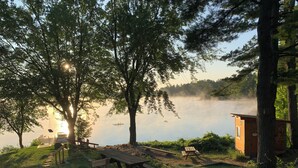 Una cabaña junto al lago con mesas de picnic y un conjunto de columpios, rodeada de árboles y vistas al lago.